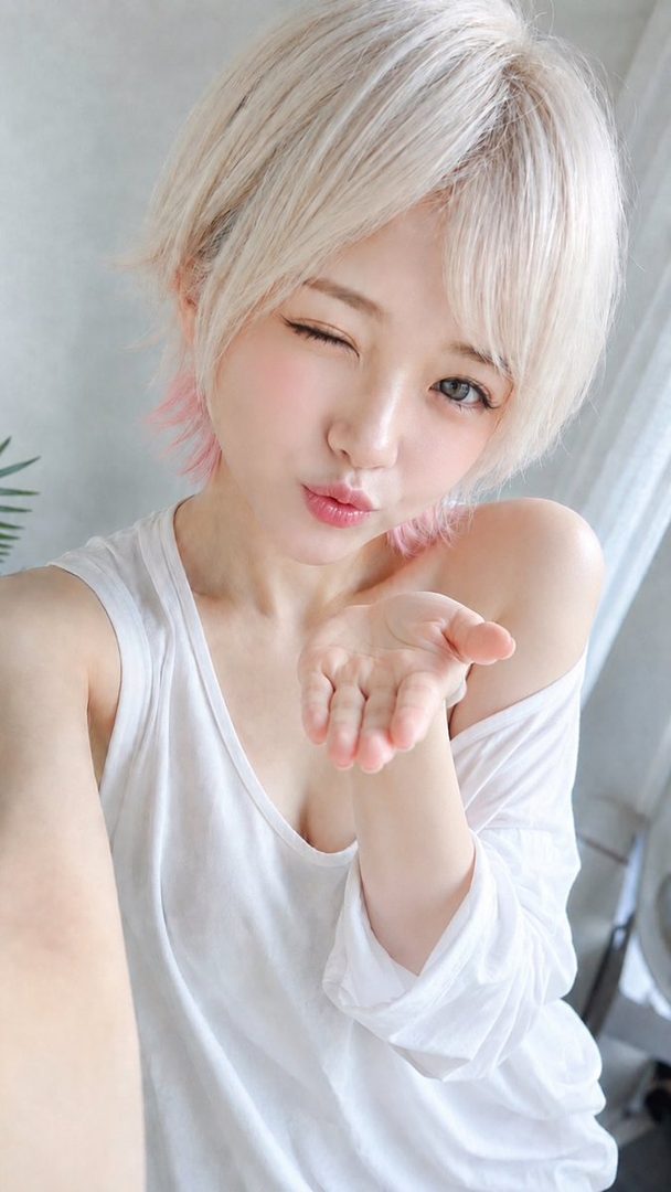 A young woman with short blonde hair and pink highlights winks and blows a kiss towards the camera, wearing a white tank top.