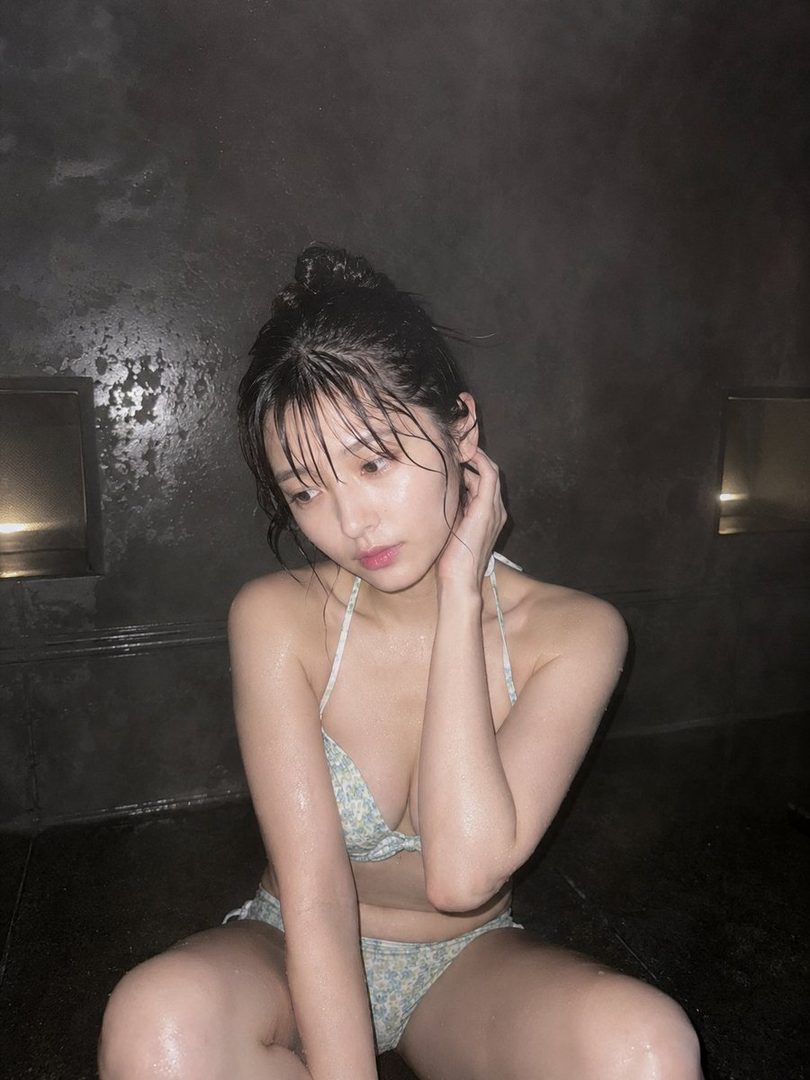 A young woman with her hair in a bun, wearing a floral bikini, sits in a steamy indoor environment, looking down.