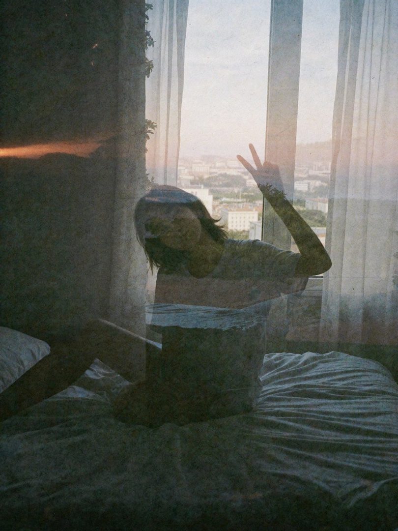 A young woman with long dark hair sits on a bed, making a peace sign with her hand, with a city view through the window.