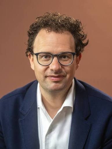 A studio portrait of a man with curly dark hair and glasses, wearing a white shirt and a dark blue suit jacket.