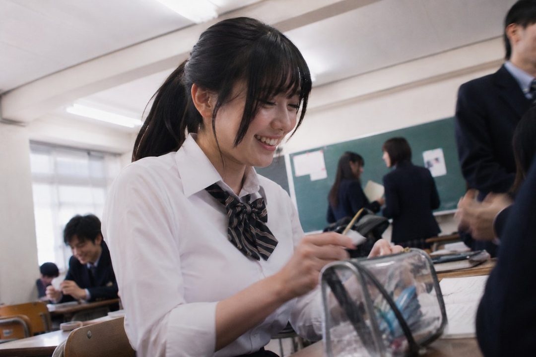 A smiling young woman in a school uniform, sitting at a desk in a classroom.