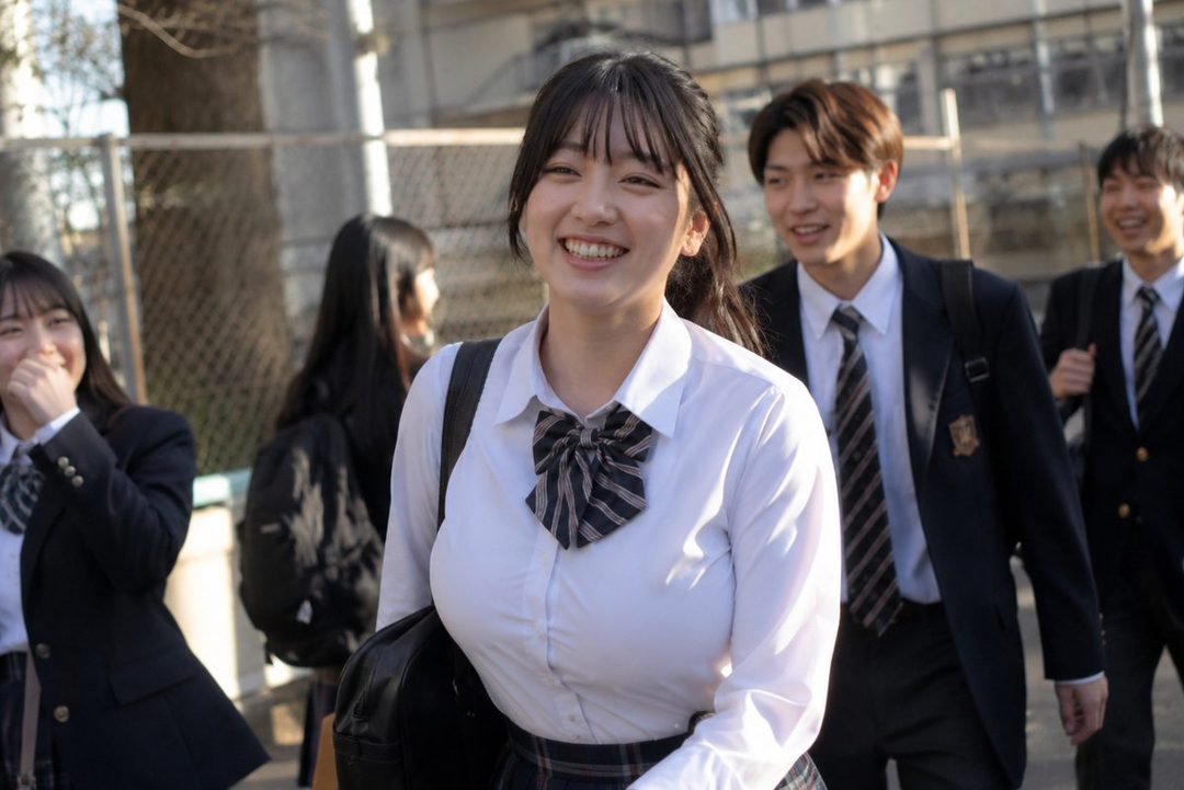 A cheerful young woman in a school uniform smiles brightly while walking with friends on a sunny day.