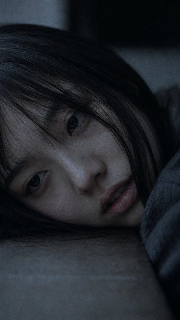 Close-up portrait of a young Japanese woman with black hair and bangs, resting her face on a surface with a distant expression, wearing a grey sweater.