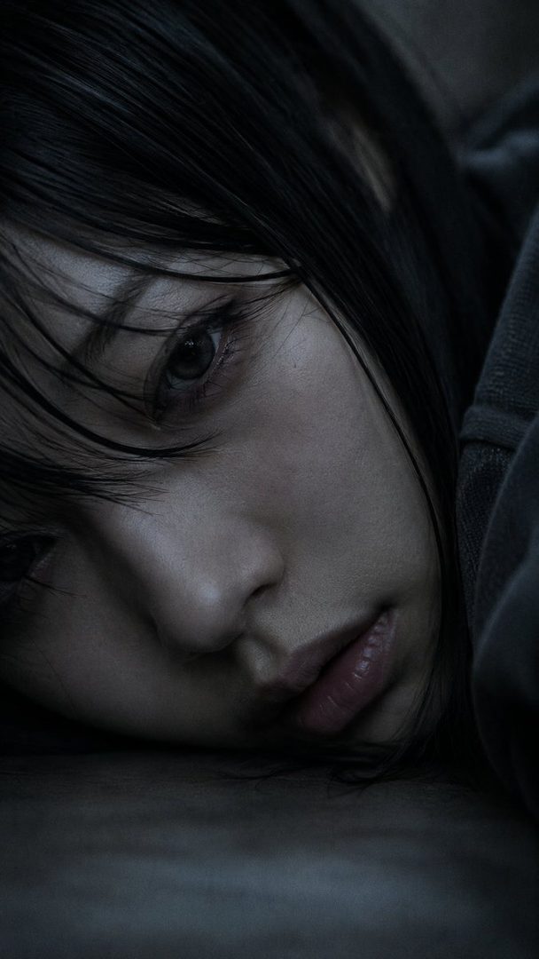 Close-up portrait of a young Japanese woman with black hair and dark eyes, resting her face on a surface with a thoughtful expression.