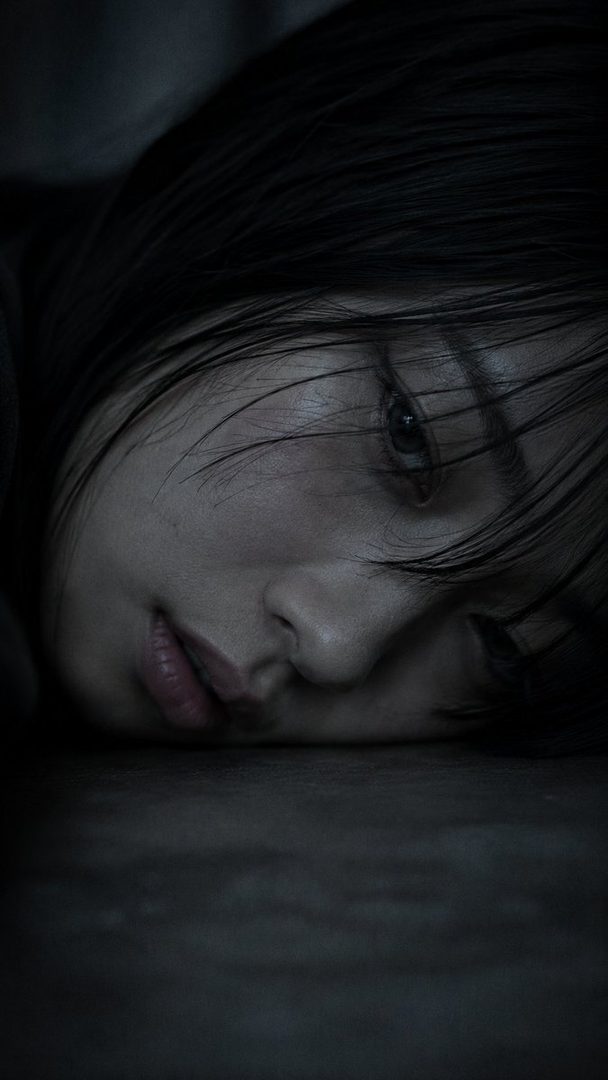 Close-up portrait of a young Japanese woman with black hair, resting her cheek on a surface, with a melancholic and absent expression.
