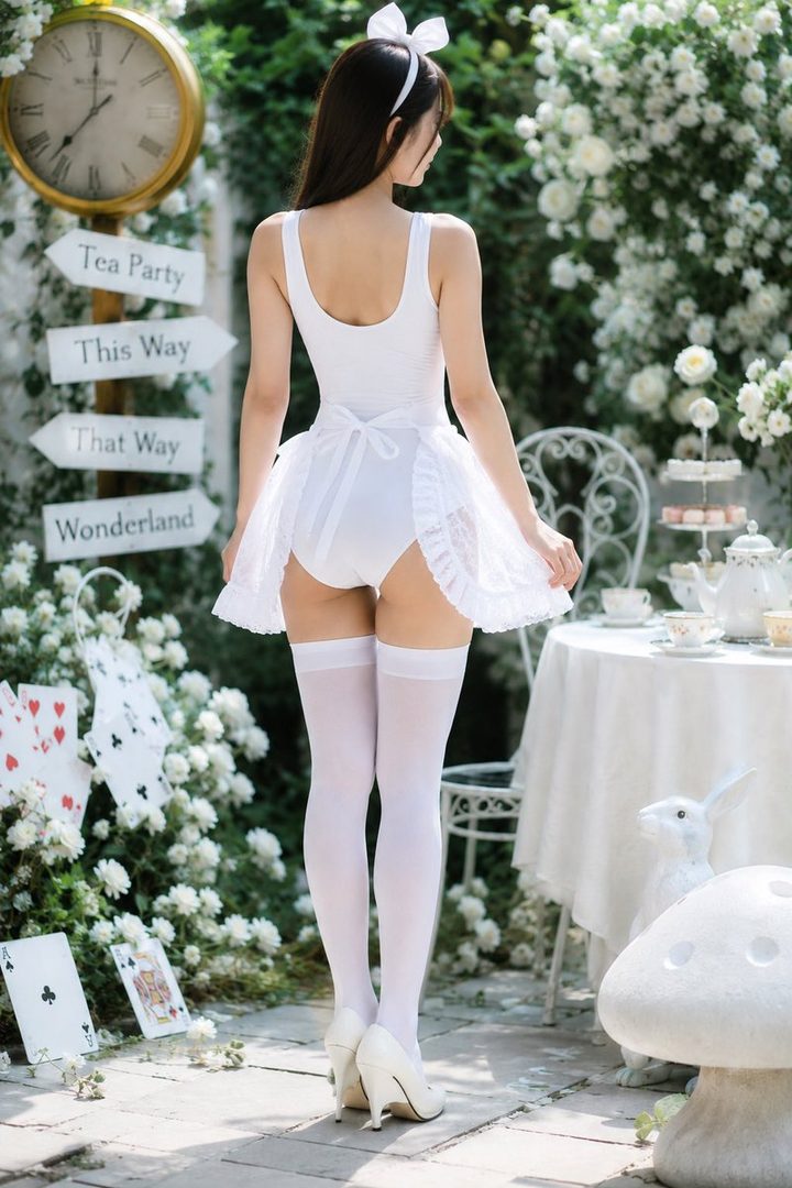A woman in a white Alice in Wonderland-themed swimsuit and white thigh-high stockings poses in a garden setting with playing cards and a tea party setup.
