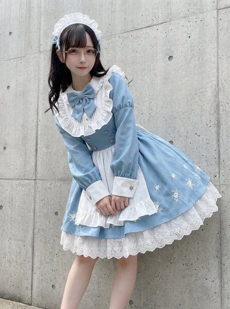 A young woman in a blue and white maid outfit with lace trim and a bow, posing outdoors against a concrete wall.