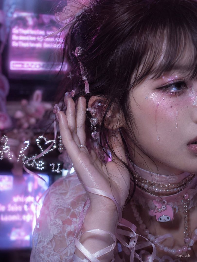 A close-up portrait of a young East Asian woman with dark hair, styled with bangs and adorned with pink ribbons and glittery makeup, wearing a lace blouse and pearl necklaces.