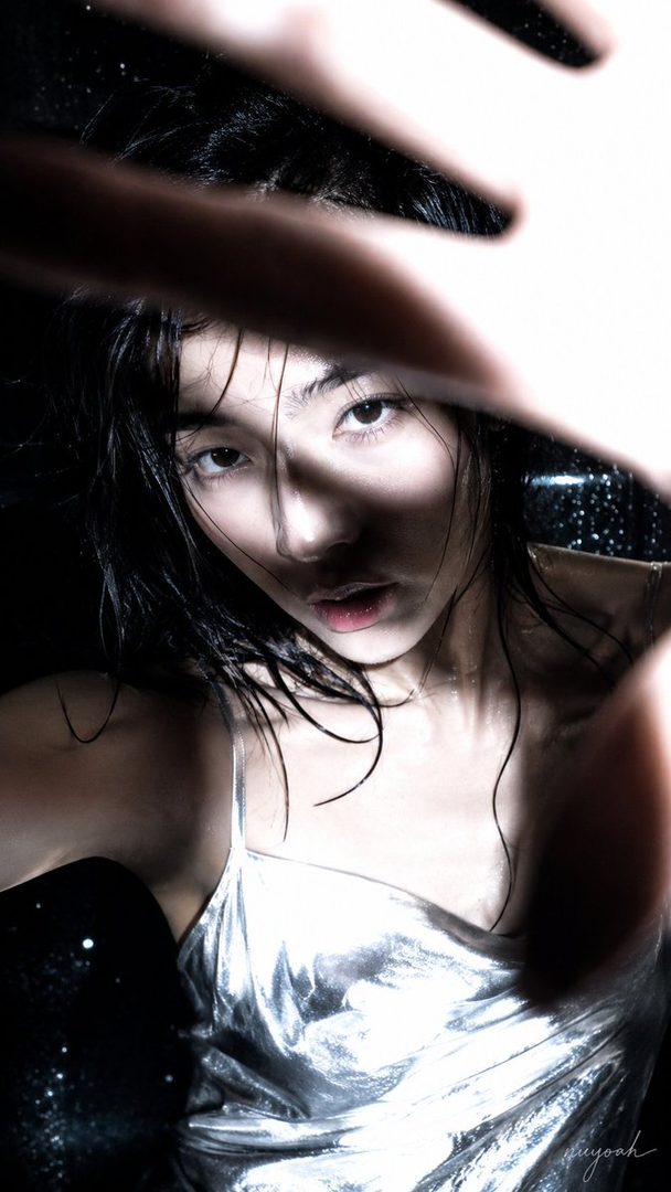 A close-up portrait of a woman with wet black hair, wearing a silver slip dress, with her hand partially obscuring her face, in a dramatic, dimly lit setting.