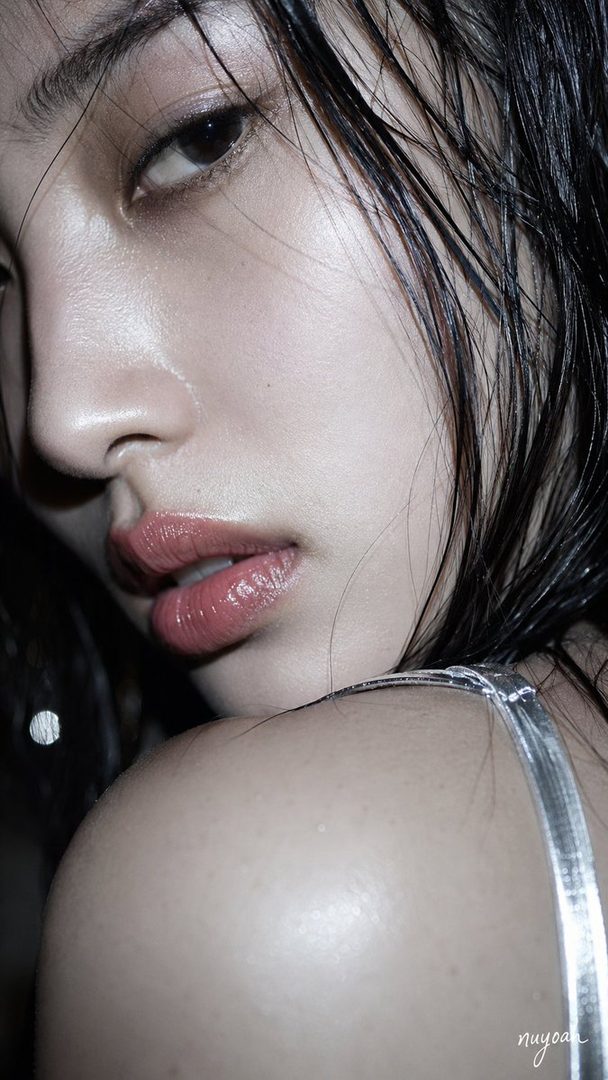 Close-up portrait of a woman with wet black hair, looking over her shoulder with intense eyes and glossy lips, wearing a silver strap.