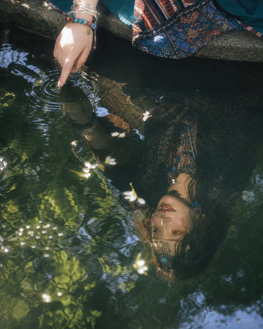 A woman with long dark hair is reflected in the water, touching the surface with her finger, creating ripples. She wears a colorful patterned top and jewelry.