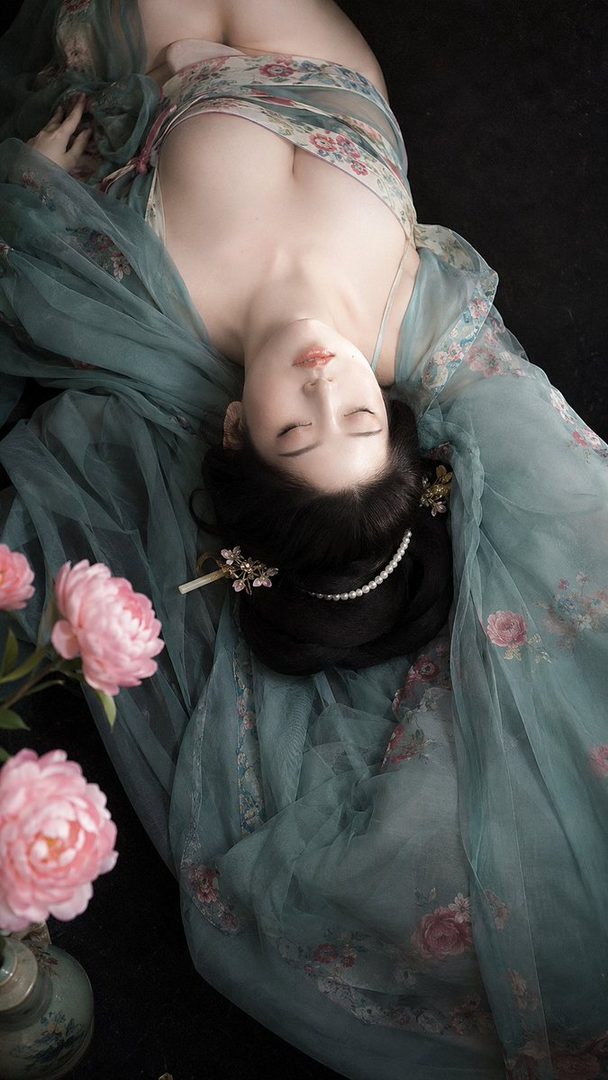 A woman with black hair styled in an updo, wearing a flowing floral dress, lies with her eyes closed, surrounded by pink peonies.