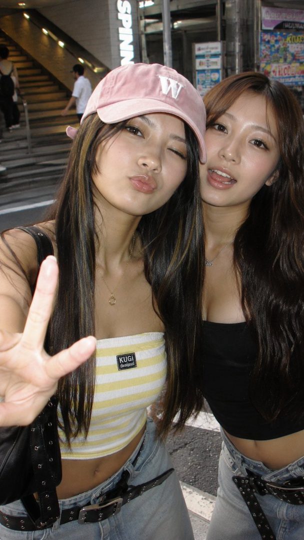 Two young East Asian women taking a selfie on a city street, one wearing a pink baseball cap and a yellow and white striped tube top, the other in a black top.