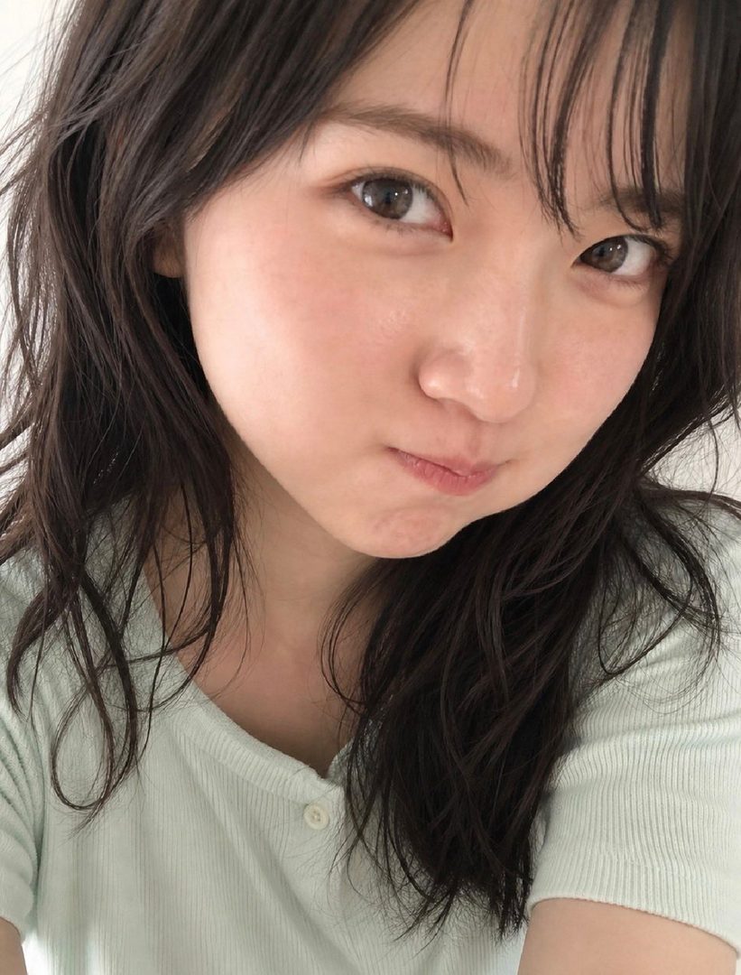 A close-up selfie of a young East Asian woman with dark, wavy hair and bangs, wearing a light green t-shirt, making a slightly pouty face.