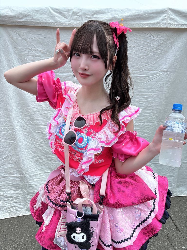 A young woman with dark hair in a ponytail, wearing a pink frilly outfit and holding a bottle of water, poses outdoors.