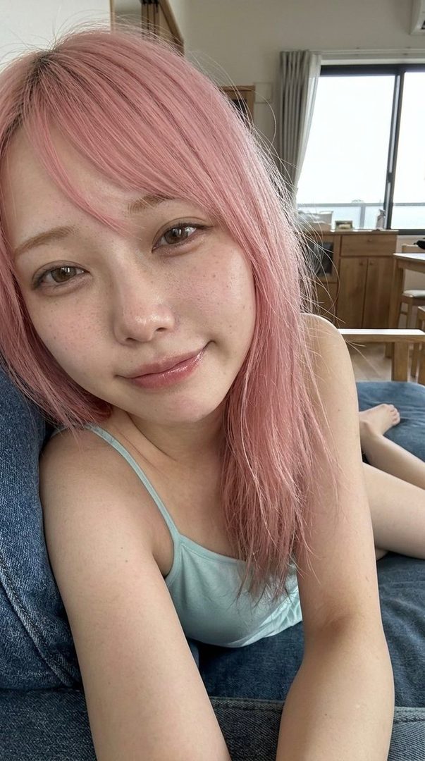 A young woman with pink hair and freckles smiles at the camera, wearing a light blue camisole and denim.