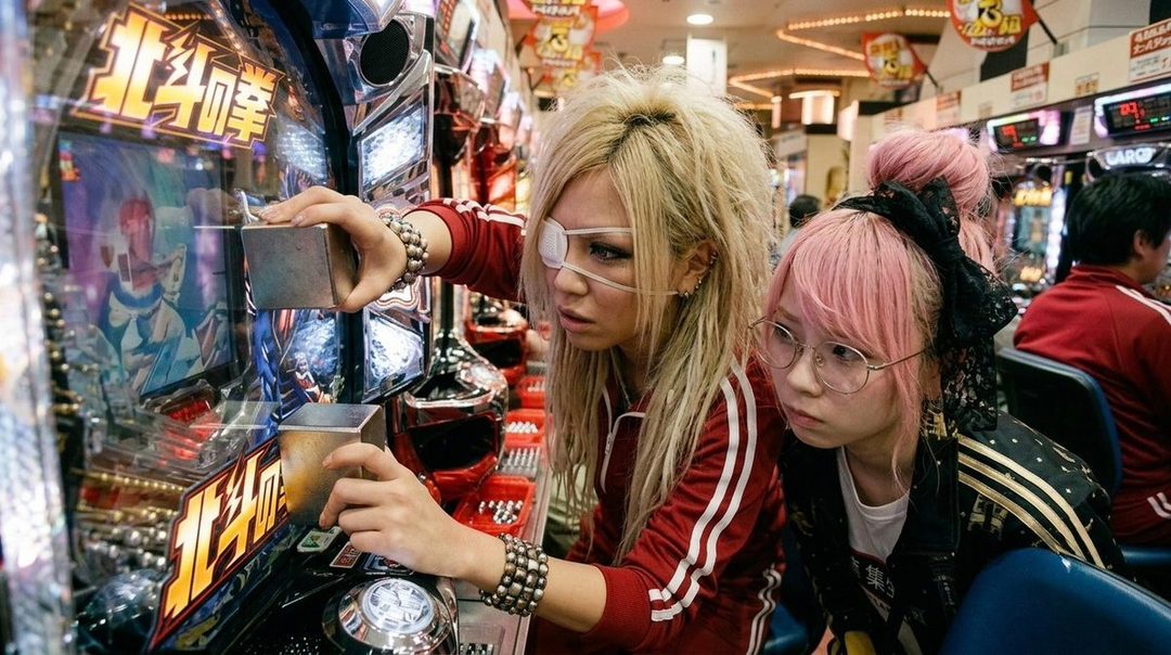 Two young women, one with blonde hair and an eye patch, and another with pink hair and glasses, are focused on playing a pachinko machine in an arcade.
