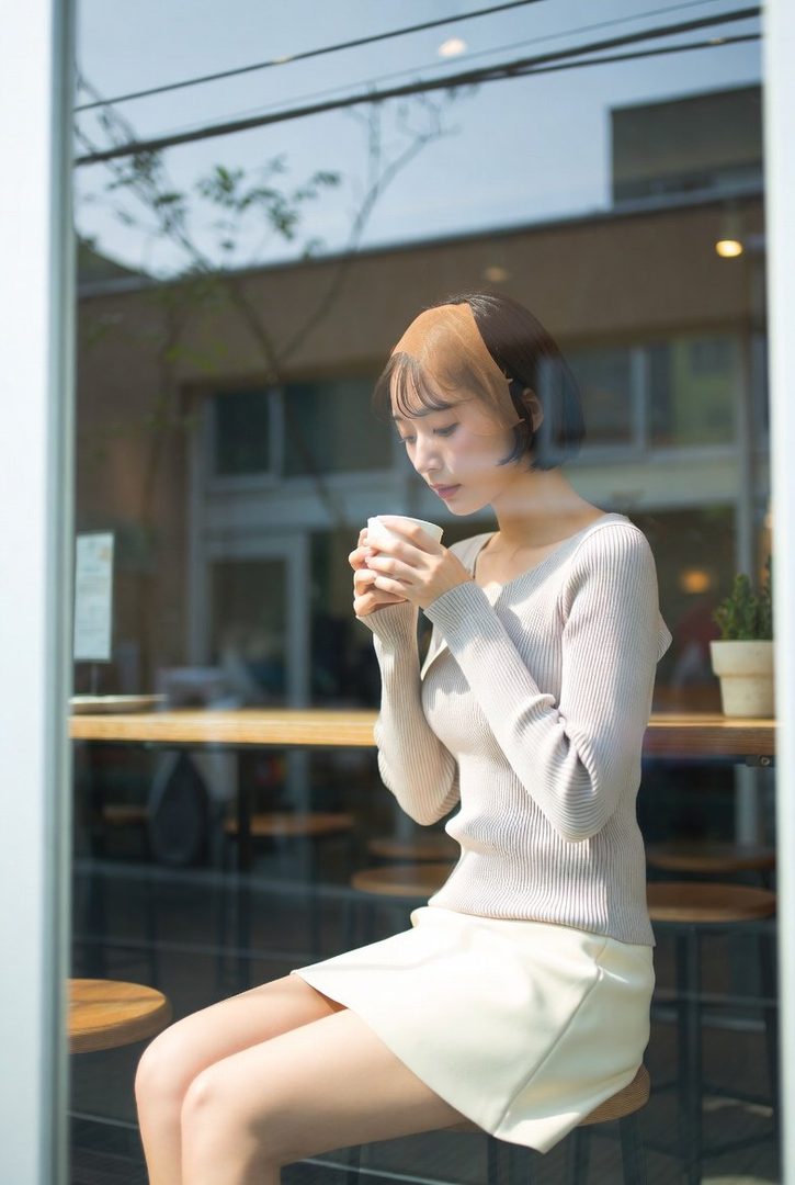 A young East Asian woman with short, two-toned hair sits by a window, holding a cup of coffee and looking down.