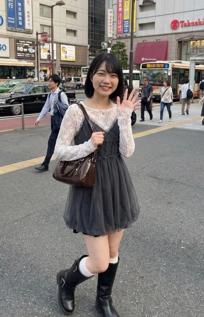 A young Japanese woman with short black hair smiles and waves while standing on a city street, wearing a dark dress over a white lace top and black boots.