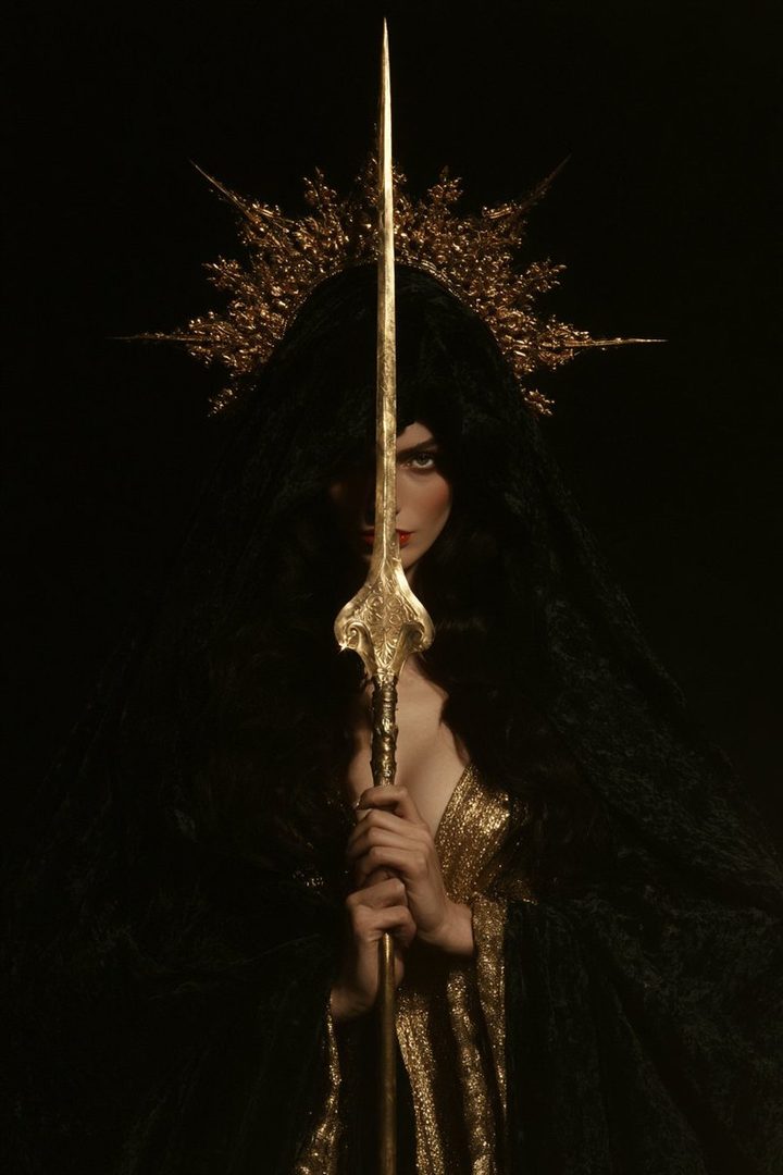 A woman with long black hair, wearing a black veil and a golden dress, holds a golden spear in front of a black background.