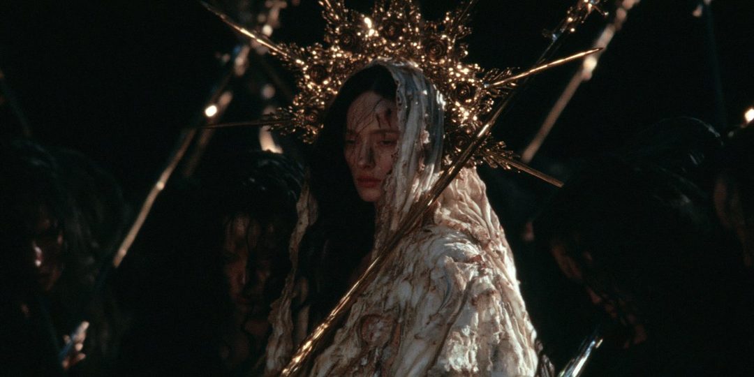 A woman with dark hair, wearing a white, textured garment and a golden halo-like headdress, looks down with a somber expression.