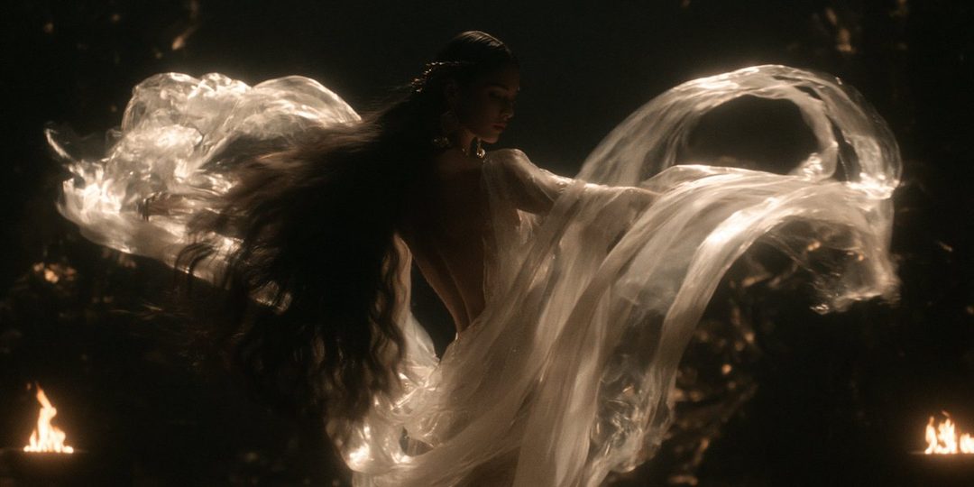 A woman with long dark hair, draped in flowing white fabric, poses dramatically in a dark setting with flickering flames.