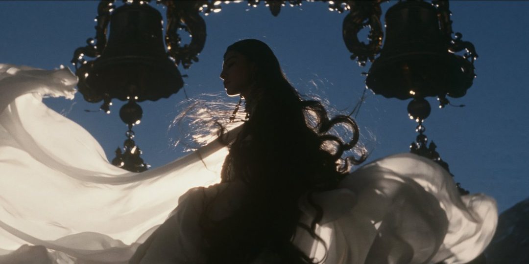 A woman with long dark hair, wearing a flowing white dress, poses dramatically under ornate bells against a clear blue sky.
