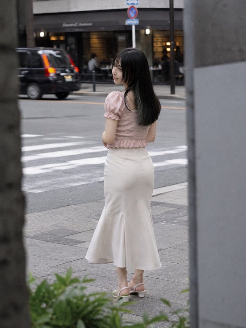 A young Japanese woman with long black hair stands on a city street, wearing a pink ruffled top and a cream-colored skirt.