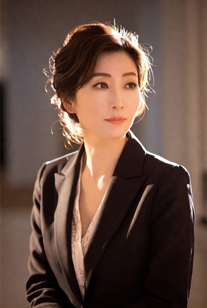 A professional portrait of an East Asian woman with short brown hair, wearing a black suit jacket over a patterned blouse, with soft lighting highlighting her features.