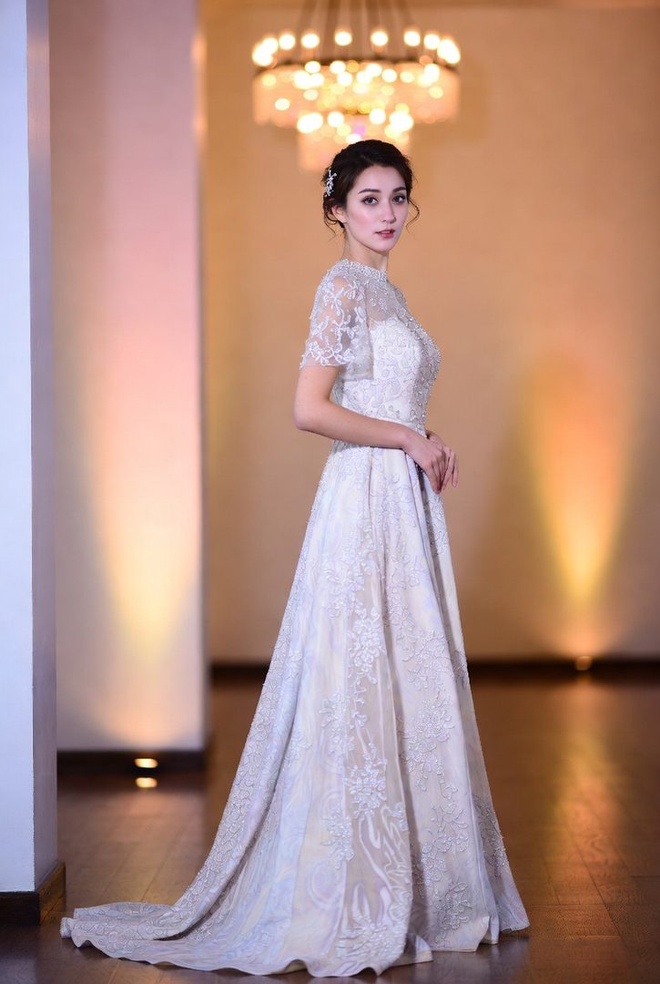 A beautiful East Asian woman in an elegant white lace wedding dress poses indoors, with soft lighting and a chandelier in the background.
