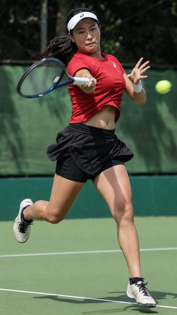 A female tennis player in a red shirt and black skirt hitting a forehand shot on a sunny day.
