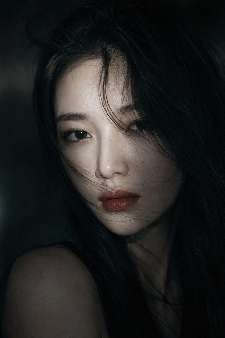 A cinematic close-up portrait of an East Asian woman with long, dark, slightly messy hair framing her face. She has fair, translucent skin, soft pink lips, and deep, expressive eyes, gazing calmly and melancholically into the camera. Dramatic low-key lighting illuminates parts of her face, creating high contrast against a dark, hazy background. A few strands of hair drift gently, adding a natural and emotional touch.