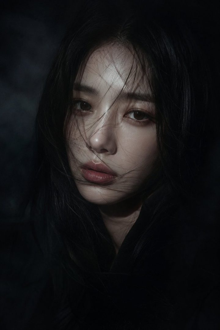 A cinematic close-up portrait of a young East Asian woman with long, dark, slightly messy hair falling across her face, looking directly at the camera with a serene and melancholic expression. Dramatic low-key lighting highlights parts of her face against a dark, hazy background, creating a moody and intimate atmosphere.