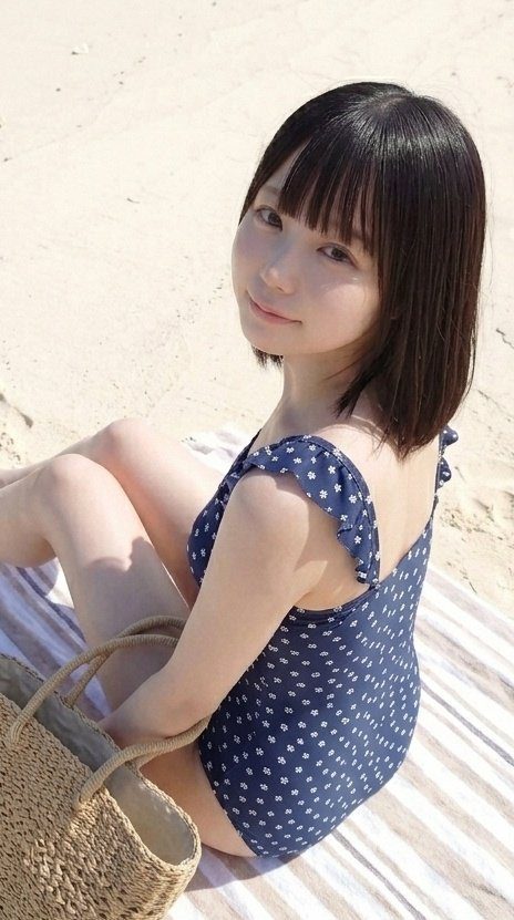 A young woman with short black hair and bangs, wearing a dark blue swimsuit with white floral patterns, sits on a striped towel on a sandy beach, looking at the camera with a gentle smile.