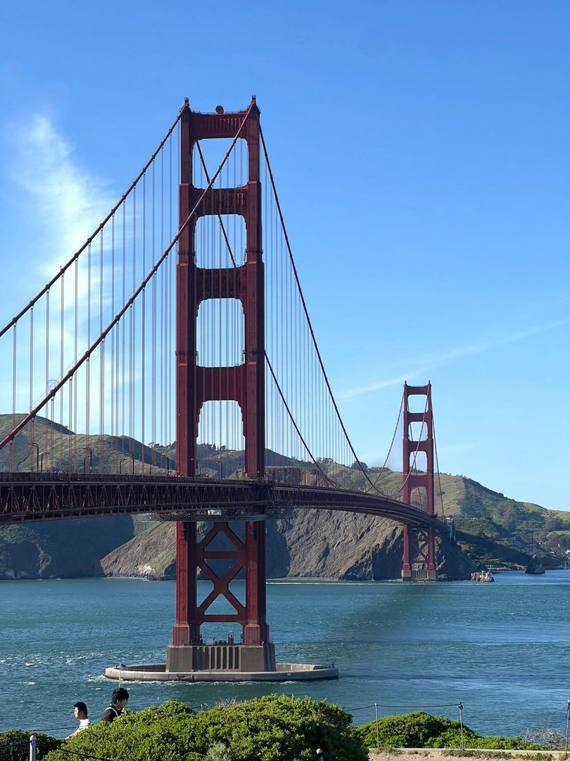 Two people stand in front of the Golden Gate Bridge on a sunny day.