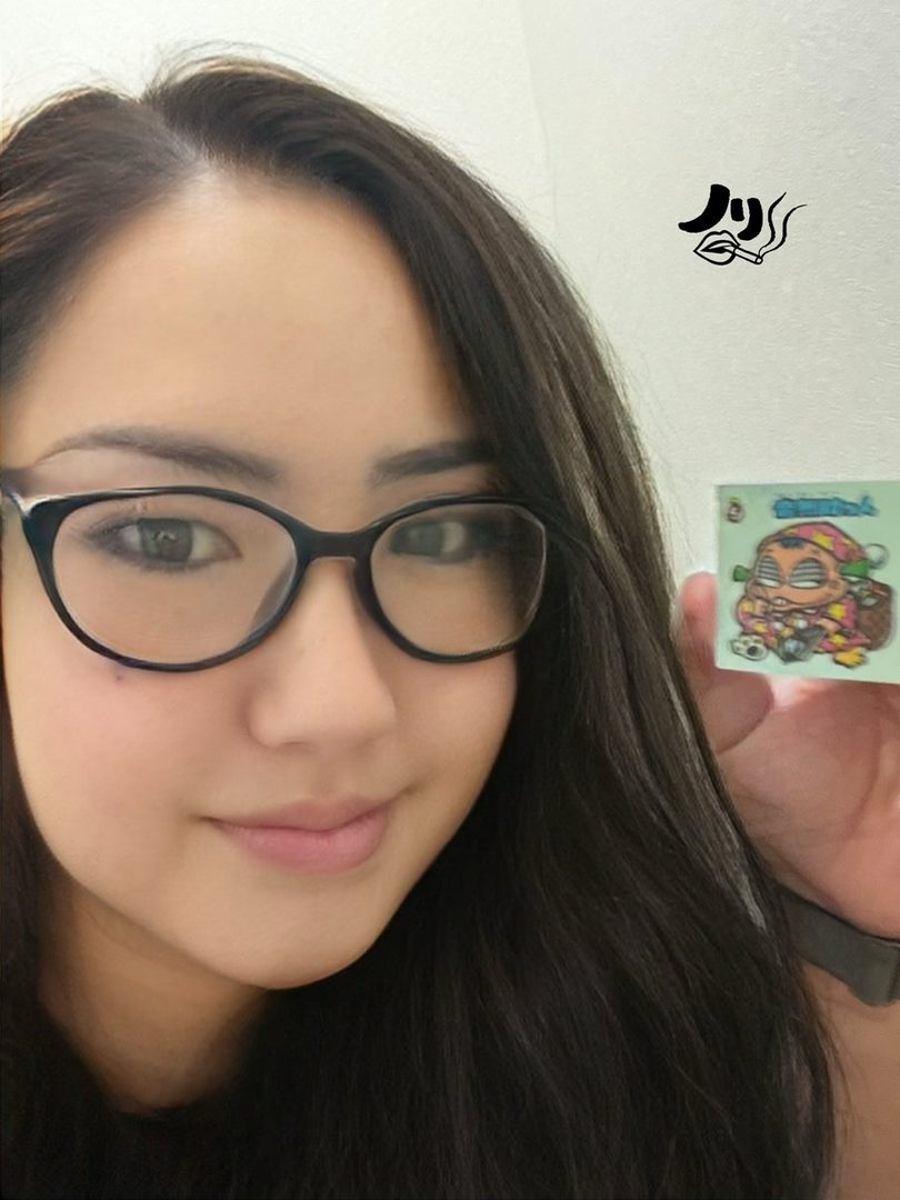 A young woman with long dark hair and glasses takes a selfie, holding up a small sticker.