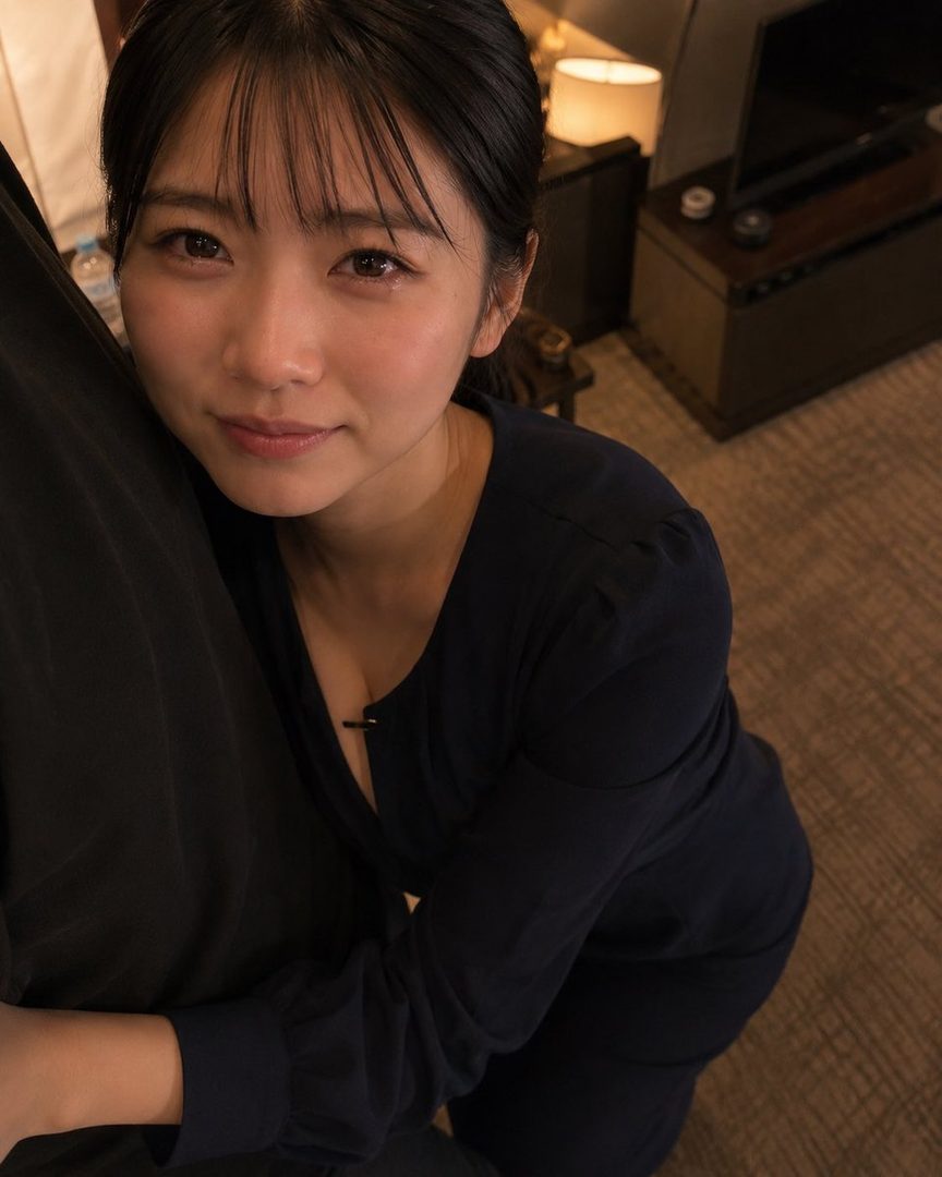 A Japanese woman with black hair and bangs, wearing a dark blue blouse, leans forward and looks at the camera with a gentle expression. The background is an indoor setting with warm lighting.