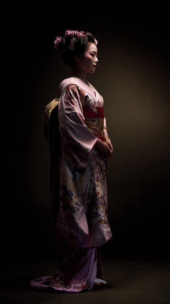 A woman in a traditional Japanese kimono stands in profile against a dark background, illuminated by dramatic lighting.