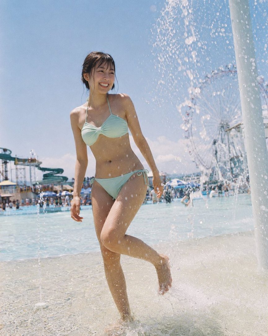 A young woman in a light blue bikini smiles as she walks through shallow water at a water park, with a Ferris wheel in the background.