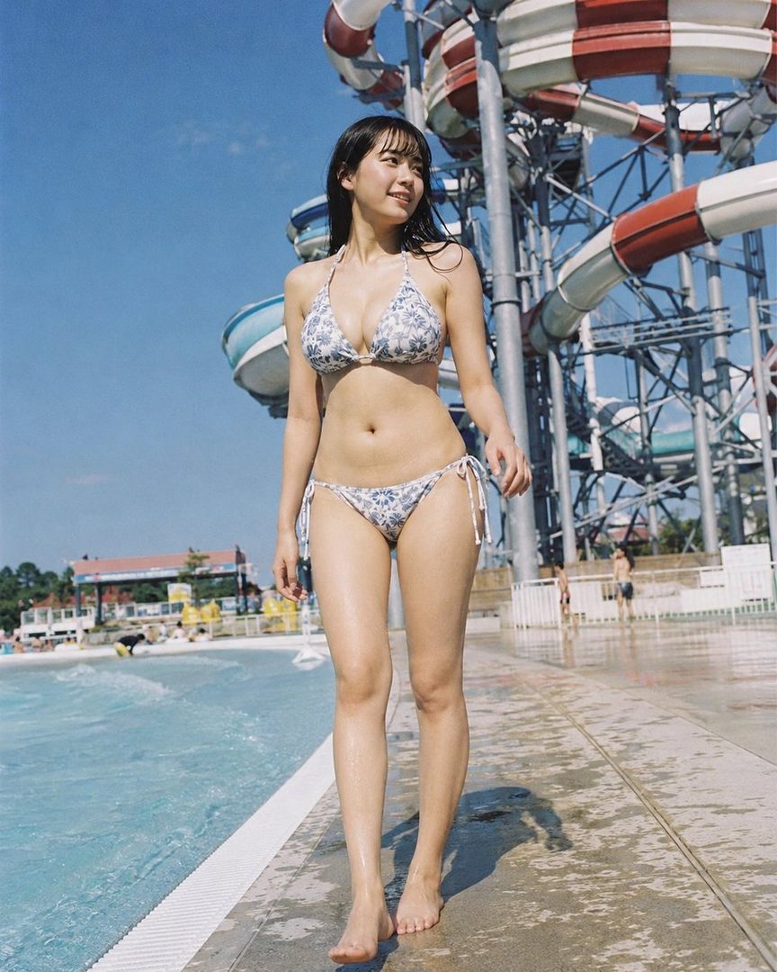 A young woman in a bikini walks by a pool at a water park, with water slides in the background.