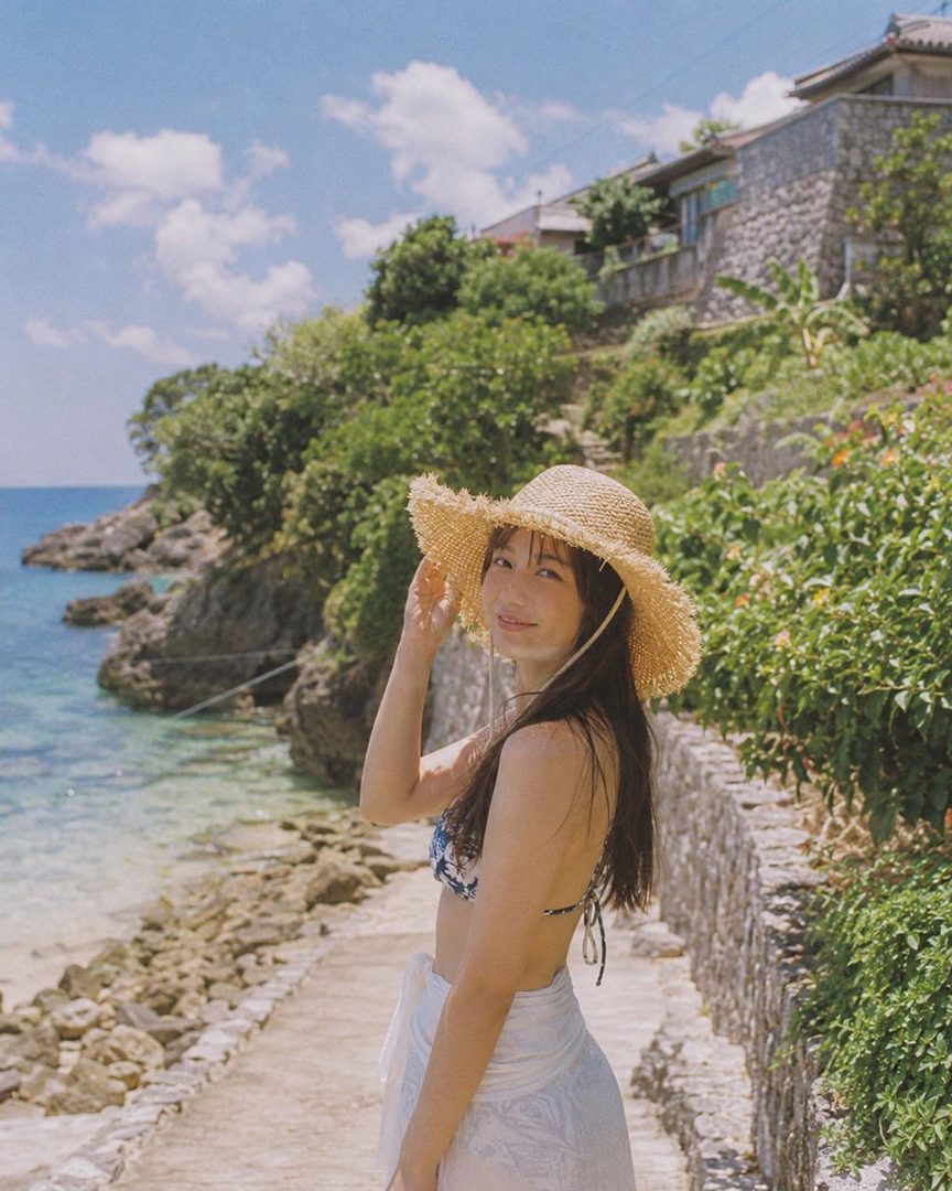 A young woman in a straw hat and bikini poses on a sunny beach path with the ocean and rocky shore in the background.