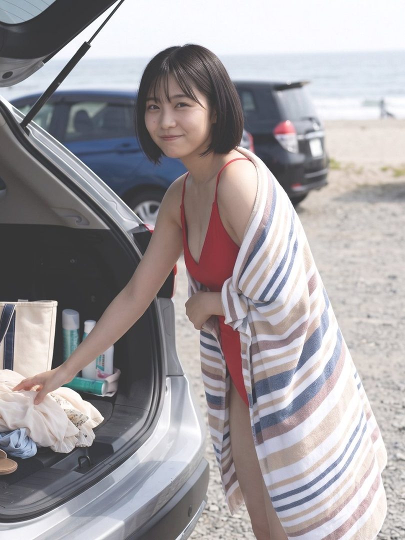 A young woman in a red swimsuit and a striped towel stands by the open trunk of a car at the beach.