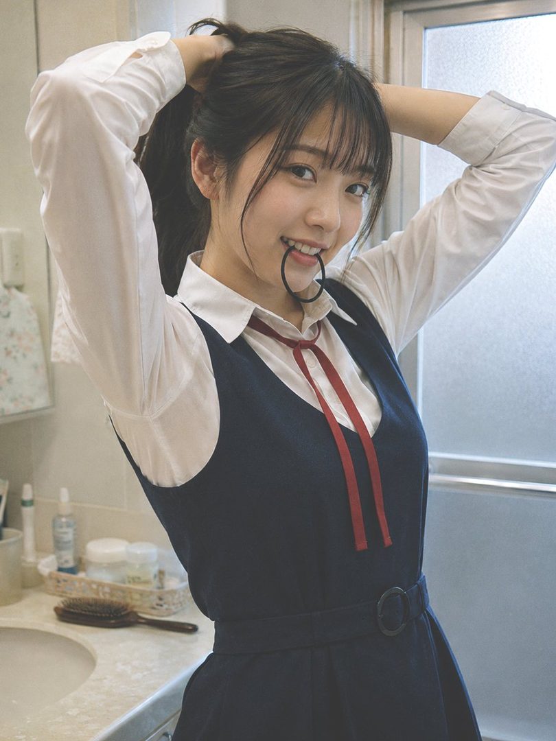 A young East Asian woman with dark hair tied in a ponytail, wearing a white shirt and a dark blue school uniform vest, smiles playfully while holding a hair tie in her mouth.