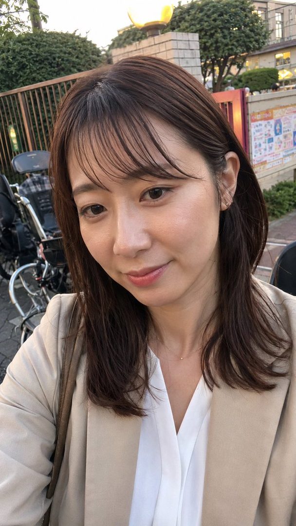 A woman with shoulder-length brown hair and bangs, wearing a beige blazer over a white blouse, takes a selfie outdoors.