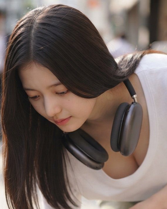 A young East Asian woman with long dark hair and headphones around her neck, looking down with a cute expression.