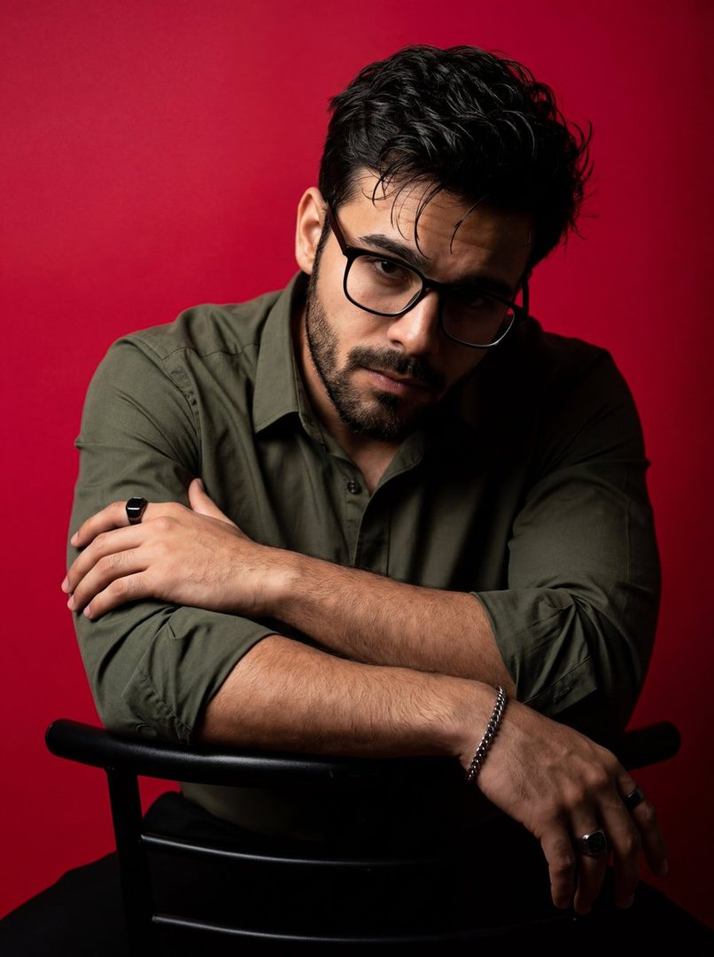 A dramatic studio portrait of a man with dark hair, beard, and glasses, wearing an olive-green shirt against a vibrant red background.