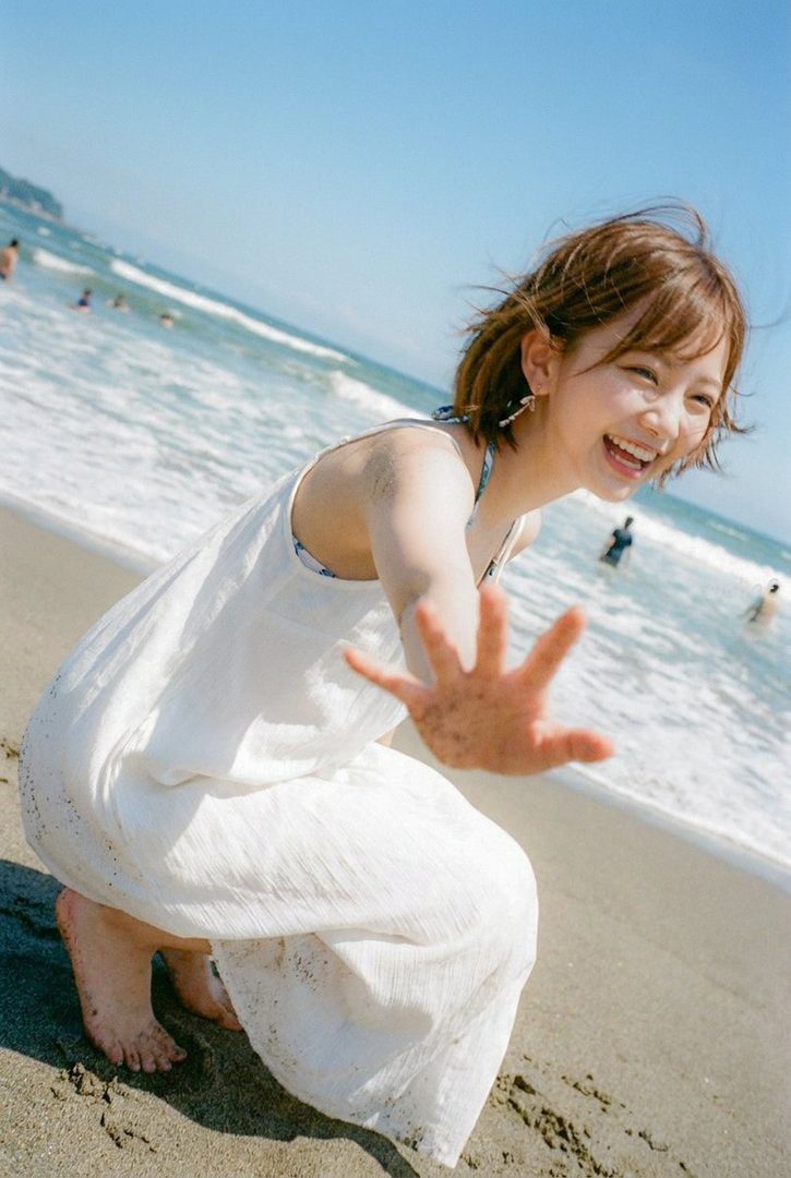 A young woman in a white dress smiles and reaches out her hand on a sunny beach with the ocean in the background.
