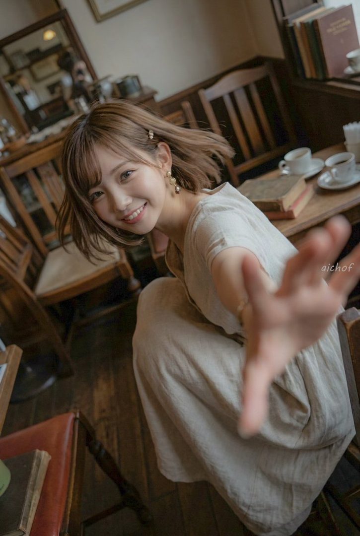 A young woman with short brown hair and bangs, wearing a light-colored dress, smiles warmly while reaching out her hand in a cozy indoor setting with wooden furniture and books.