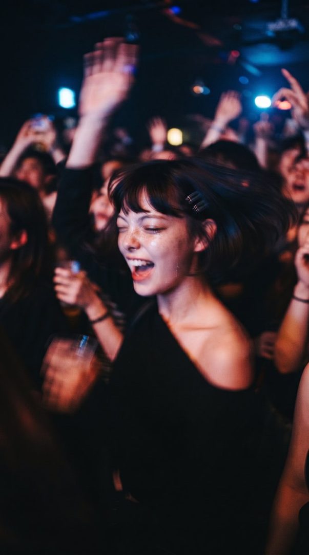 A young woman with short dark hair and bangs, wearing a black off-the-shoulder top, smiles with her eyes closed and mouth open while dancing at a concert or club.