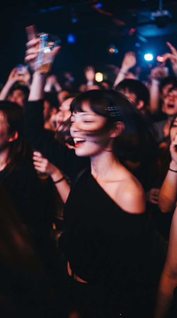 A young woman with short black hair and bangs, wearing a black off-the-shoulder top, smiles and laughs while dancing in a crowded, dimly lit club with colorful lights.